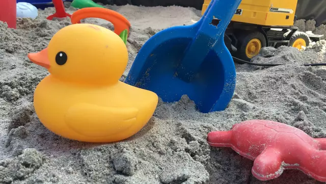 Eine gelbe Plastikente mit Schaufel und weiterem Sandspielzeug im Sandkasten.