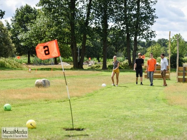 Zielfahne im fordergrund mit orangenem Wimpel auf grüner Wiese im Hintergrund Fußballgolf spielende Menschen